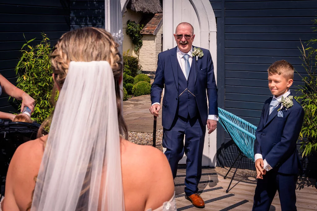 A smiling father and a young page boy in navy suits look toward a bride wearing a veil during an outdoor summer wedding ceremony.