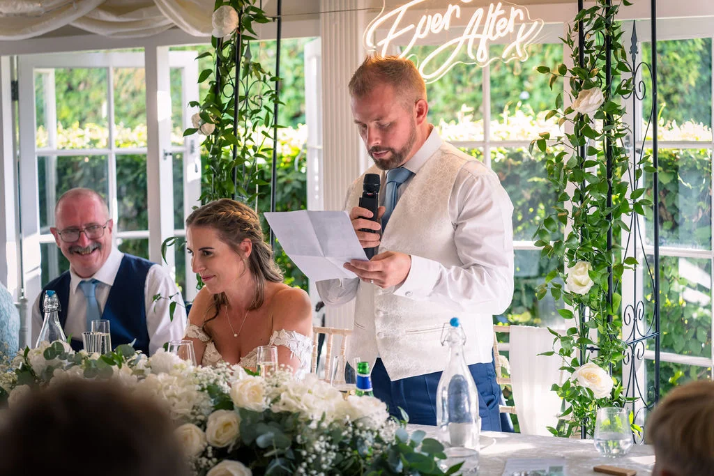 A groom delivers his speech while reading notes at the top table, with the smiling bride and an older guest seated beside him against a backdrop of white floral arches and a glowing neon sign.