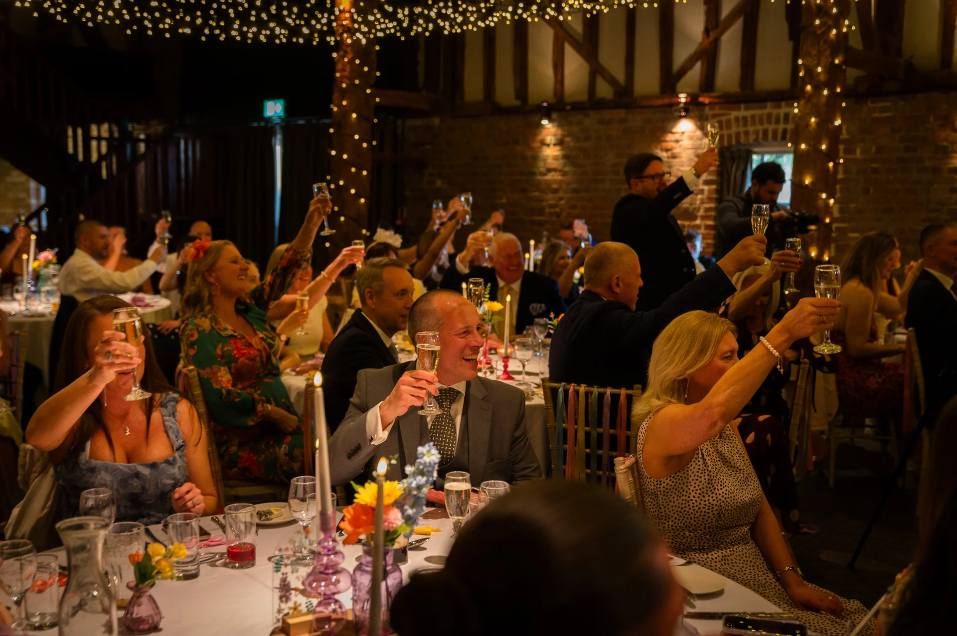 Wedding guests in a rustic barn venue with a fairy light canopy enthusiastically raise glasses of champagne during the speeches.