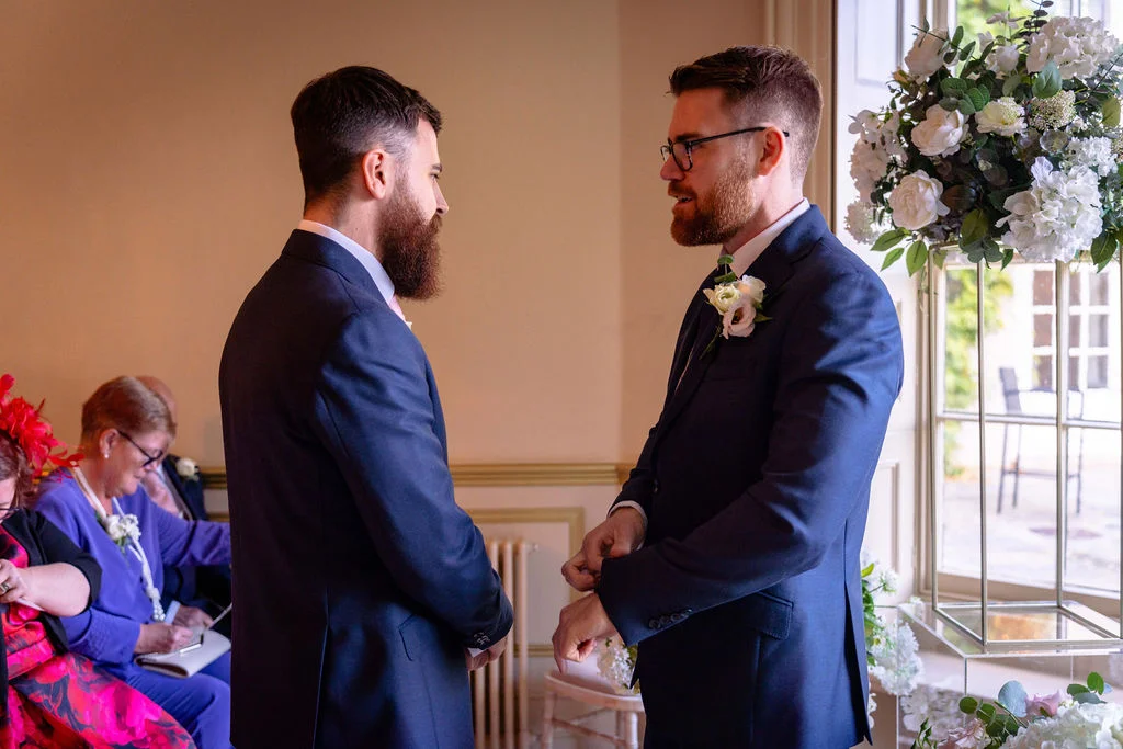 Two grooms in matching navy suits stand facing each other inside a light-filled room during their wedding ceremony, with guests seated in the background.
