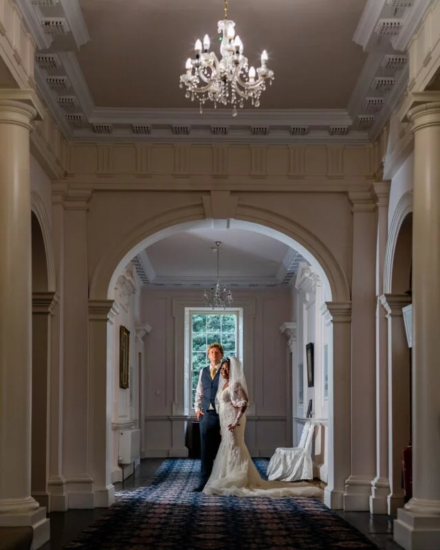 An interracial bride and groom pose elegantly inside a grand historic manor house, framed perfectly by classical stone archways and towering columns beneath an ornate chandelier.