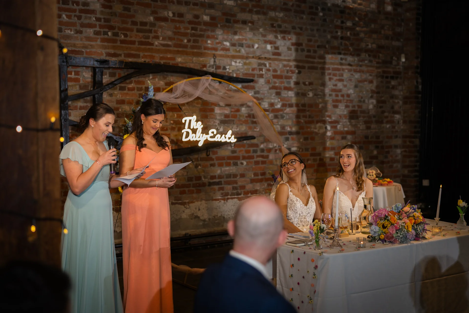 Two bridesmaids deliver speeches at the top table while the smiling couple and guests look on in a rustic brick venue.