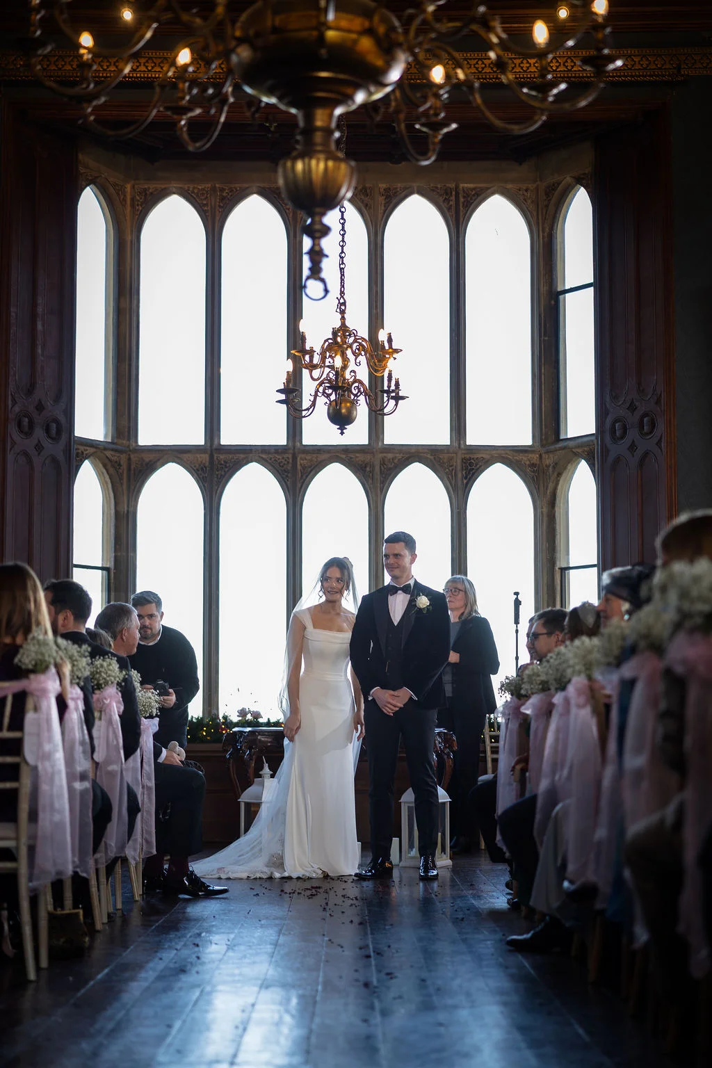 A bride and groom stand together at the front of a grand, historic hall with large arched windows and a striking chandelier during their wedding ceremony.