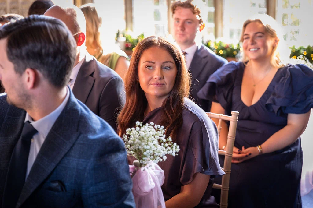 A candid photograph of wedding guests seated during a ceremony, with warm natural light illuminating the scene from behind.