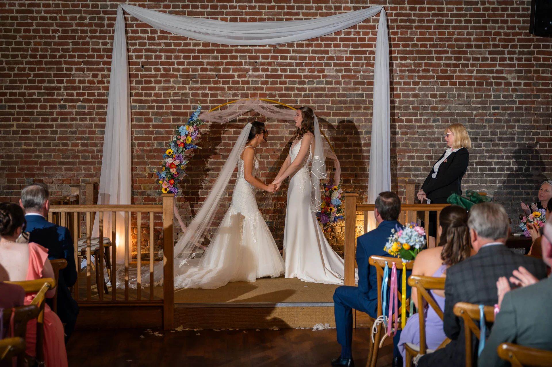 Two smiling brides hold hands facing each other during their indoor wedding ceremony before an exposed brick wall decorated with a floral hoop and white fabric draping.