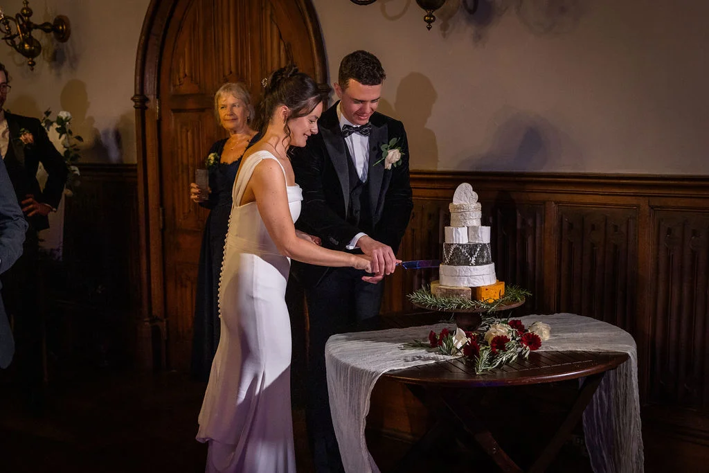 The bride and groom smile as they cut into a tiered cheese wheel cake together during their evening reception in a wood-panelled hall.
