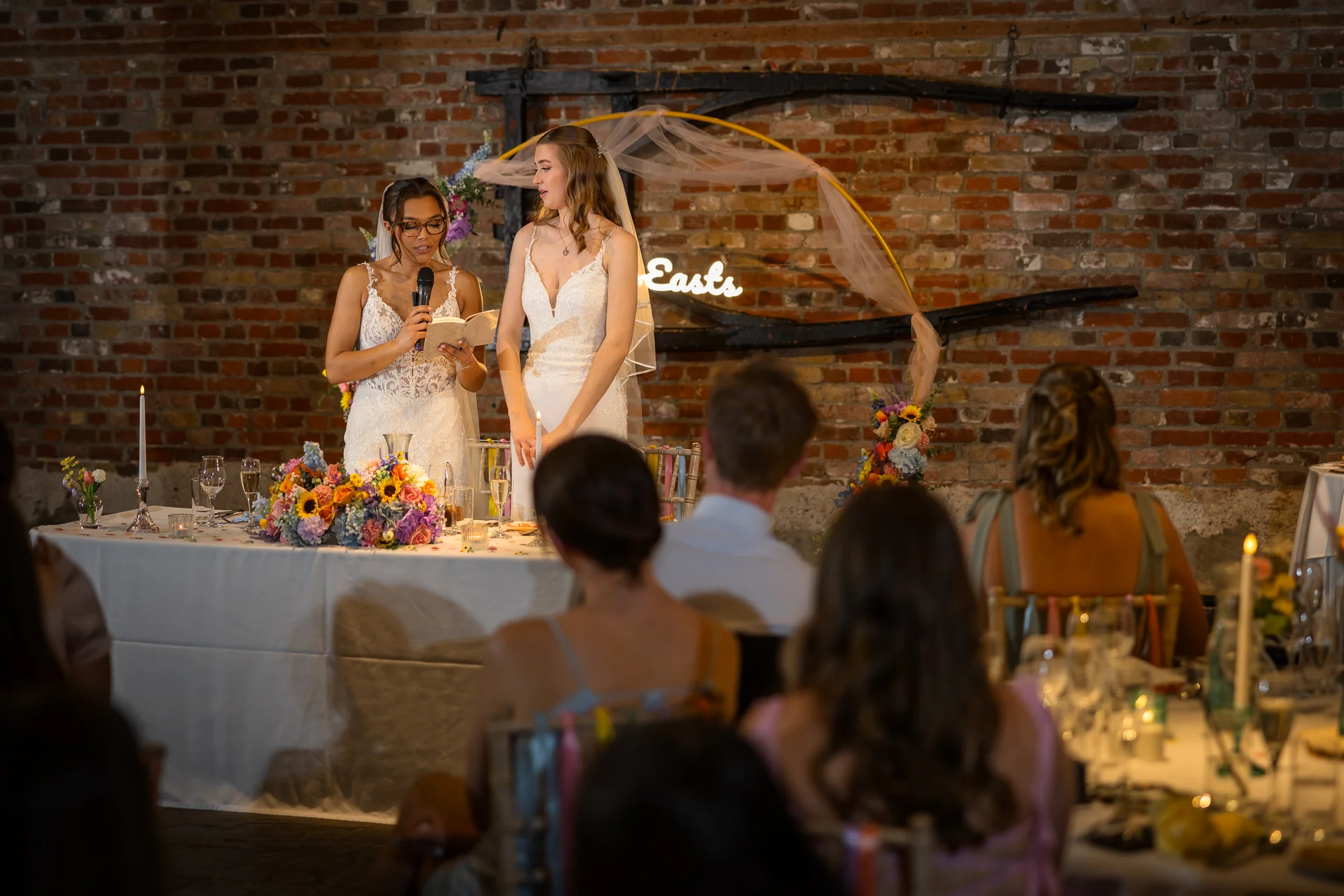 Two brides deliver a speech at the top table in a rustic brick barn venue decorated with an illuminated sign and floral arrangements.