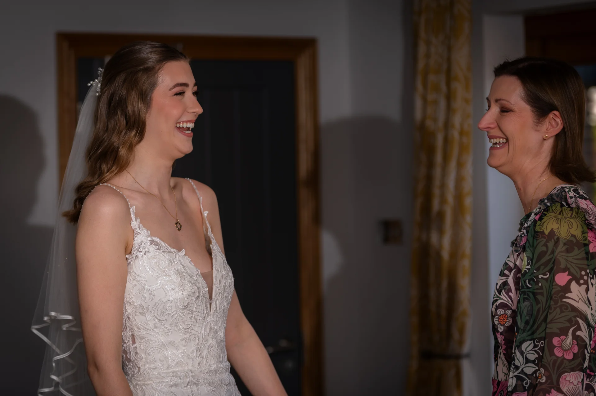 A glowing bride in a lace gown and veil laughs joyfully while sharing a tender moment with her mother indoors.