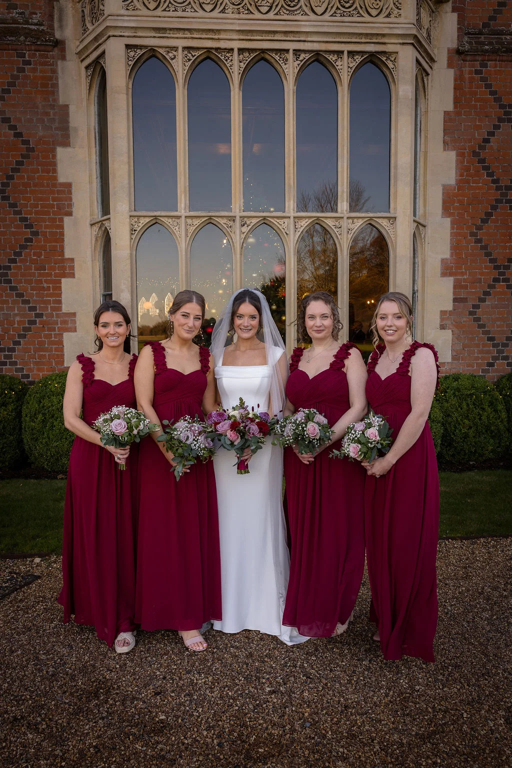 A beautiful bride in a sleek white gown and veil stands in the centre of her four bridesmaids, who wear matching burgundy dresses with floral strap details, outside a grand historic manor house.