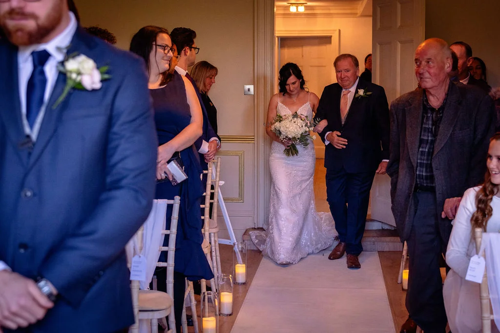 A smiling bride walks down the aisle arm in arm with her father as guests look on during the wedding ceremony.