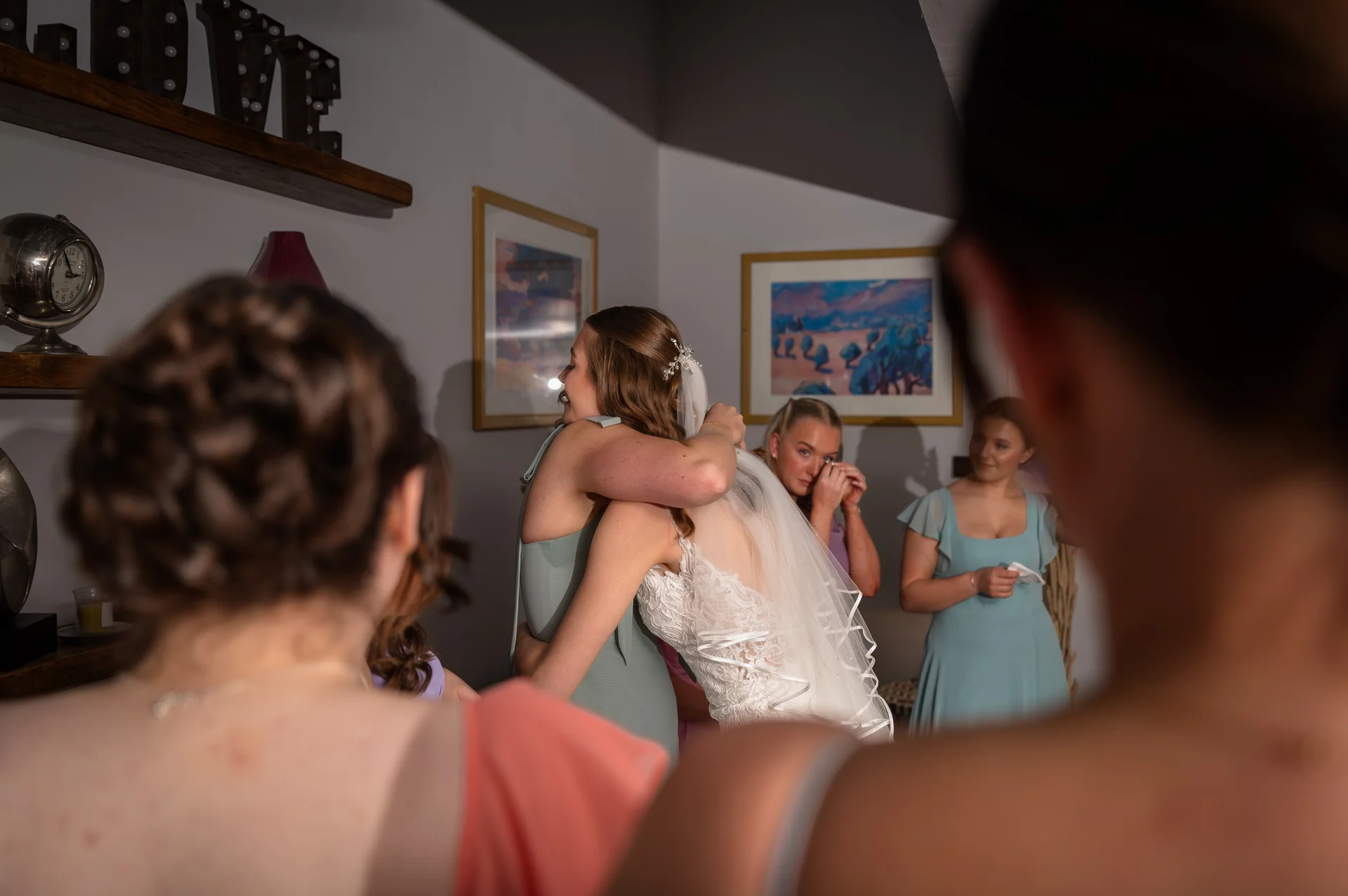 An emotional bride shares a warm hug with a bridesmaid in sage green during morning preparations while other bridal party members watch on.