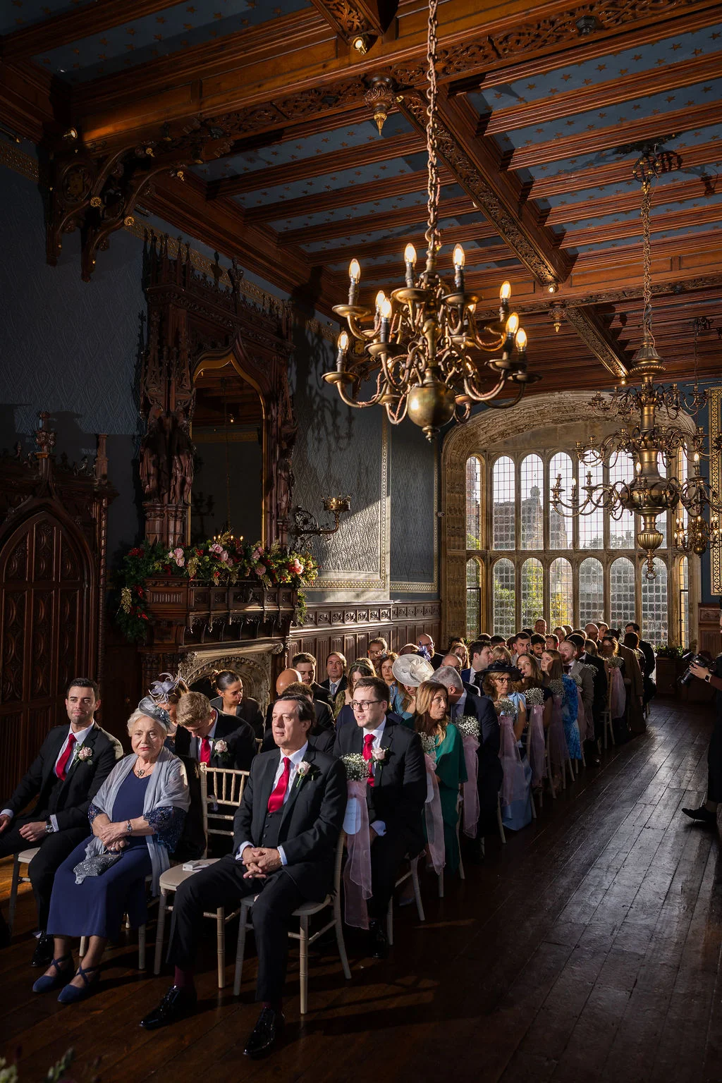 Guests are seated in a grand wood-panelled hall with a vaulted ceiling and large arched windows for a traditional wedding ceremony.