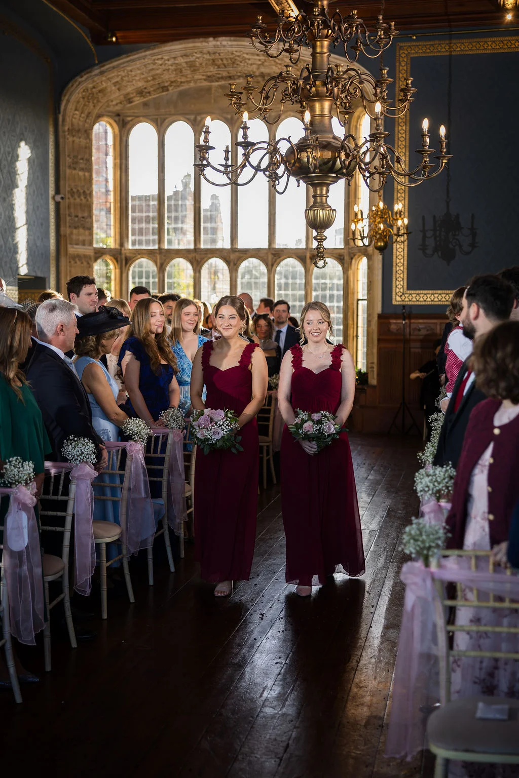 Two smiling bridesmaids in matching burgundy dresses walk down the aisle holding bouquets during a ceremony in a grand country house featuring large arched windows and a statement chandelier.