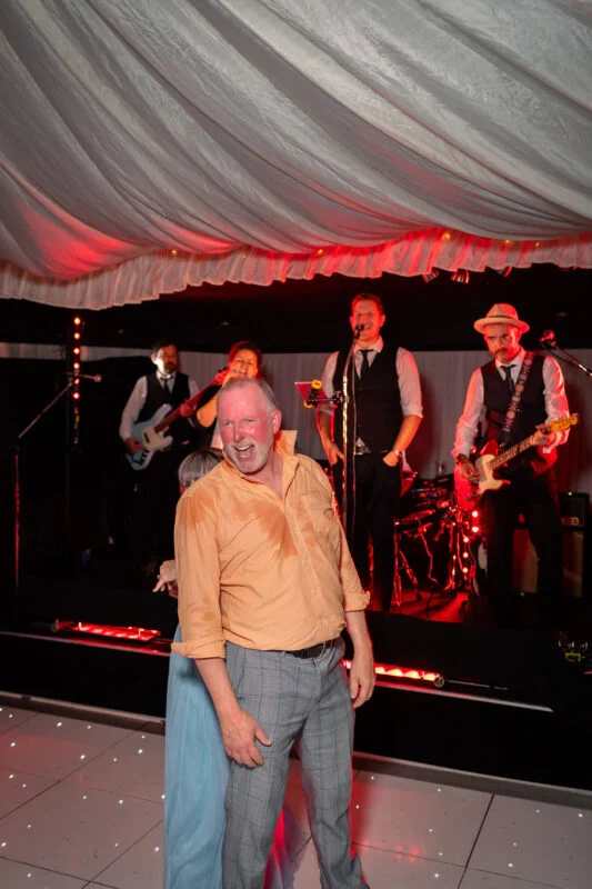 A guest laughing joyfully on a lit dance floor with a live band performing on stage inside a draped marquee.