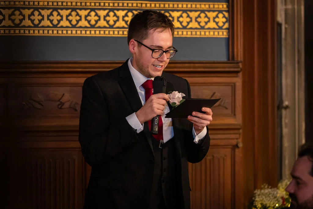 A groom wearing glasses and a black three-piece suit delivers a speech while holding a microphone and speech notes.