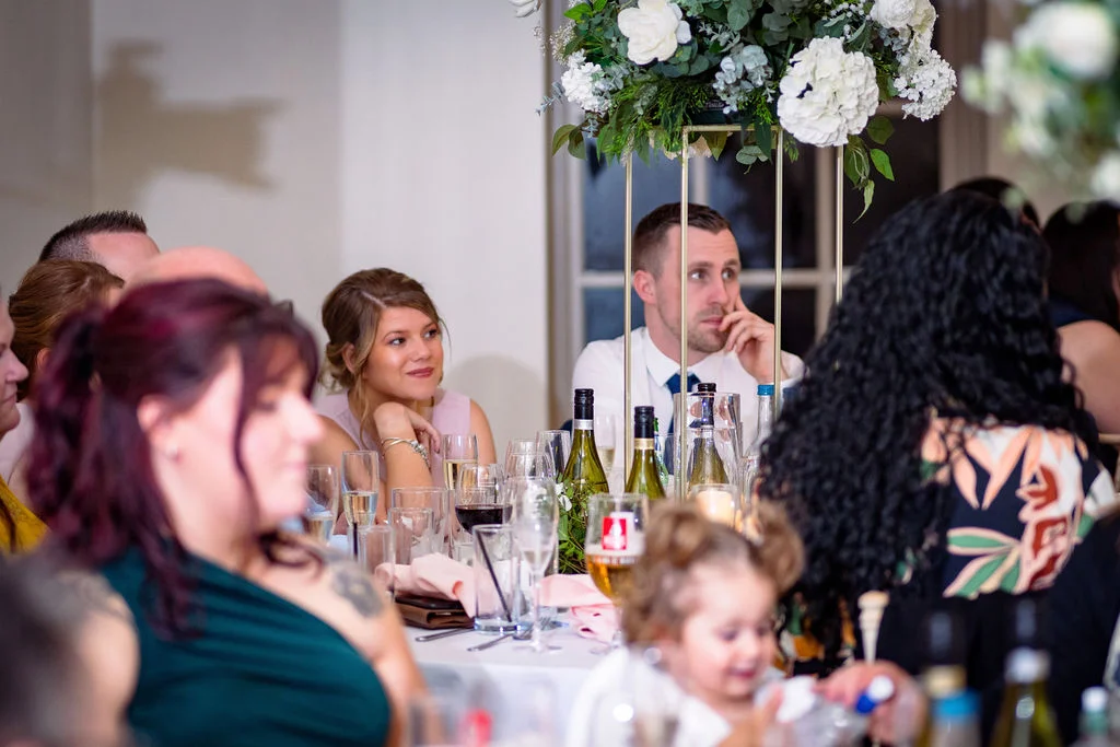 Wedding guests attentively watch the speeches during the wedding breakfast, seated at a beautifully set table adorned with tall floral centrepieces and wine glasses.