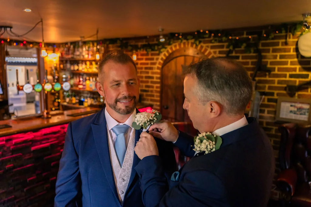 A groom in a blue suit and waistcoat has his boutonniere adjusted by his partner inside a cosy pub venue featuring exposed brickwork.