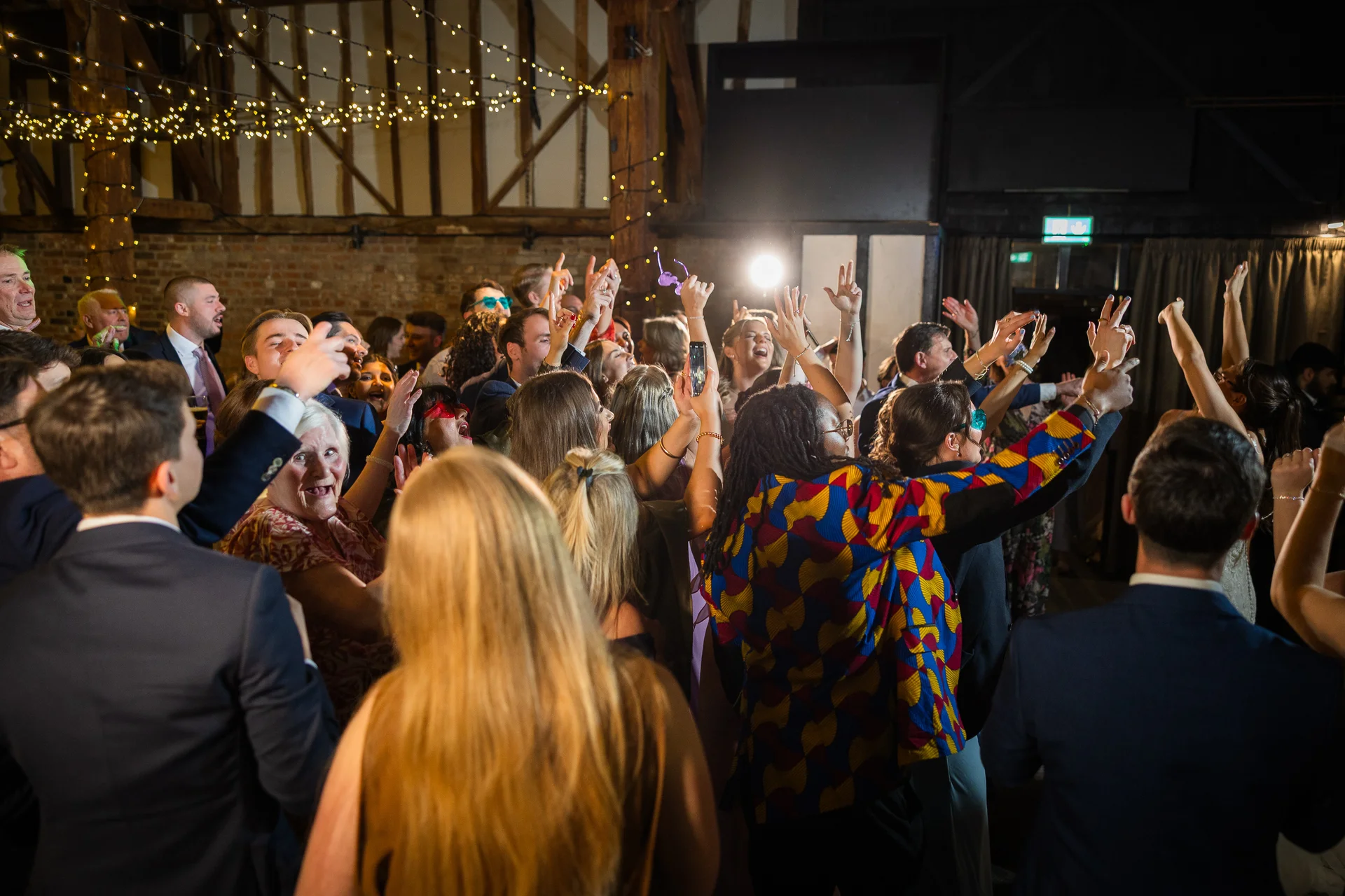 An energetic crowd of wedding guests celebrates with hands raised high on a packed dance floor under fairy lights inside a rustic oak-framed barn.
