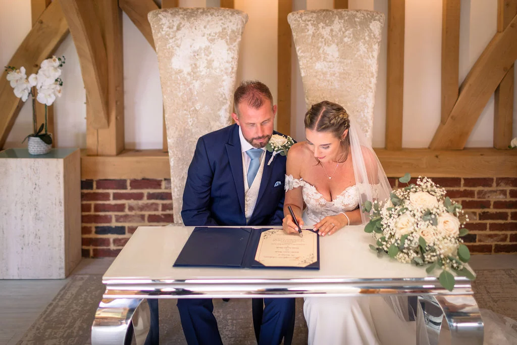 The bride and groom sit side by side at a glossy white signing table inside a rustic oak-framed barn with exposed brick walls, signing their wedding certificate.