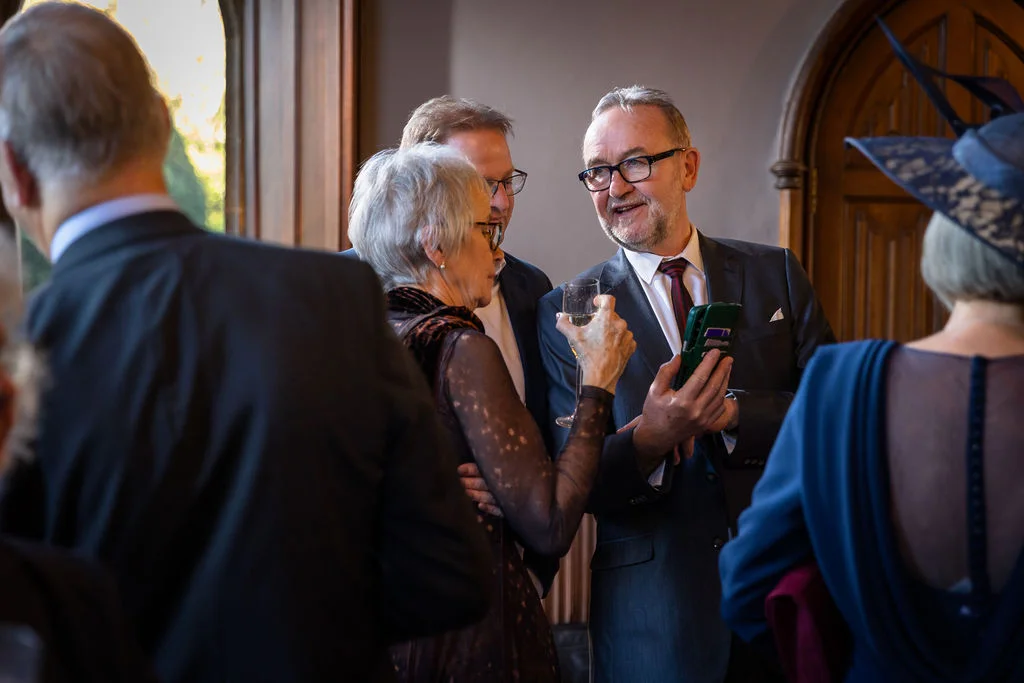 Wedding guests mingling and chatting happily during a drinks reception, with a man holding a phone and a glass of champagne.