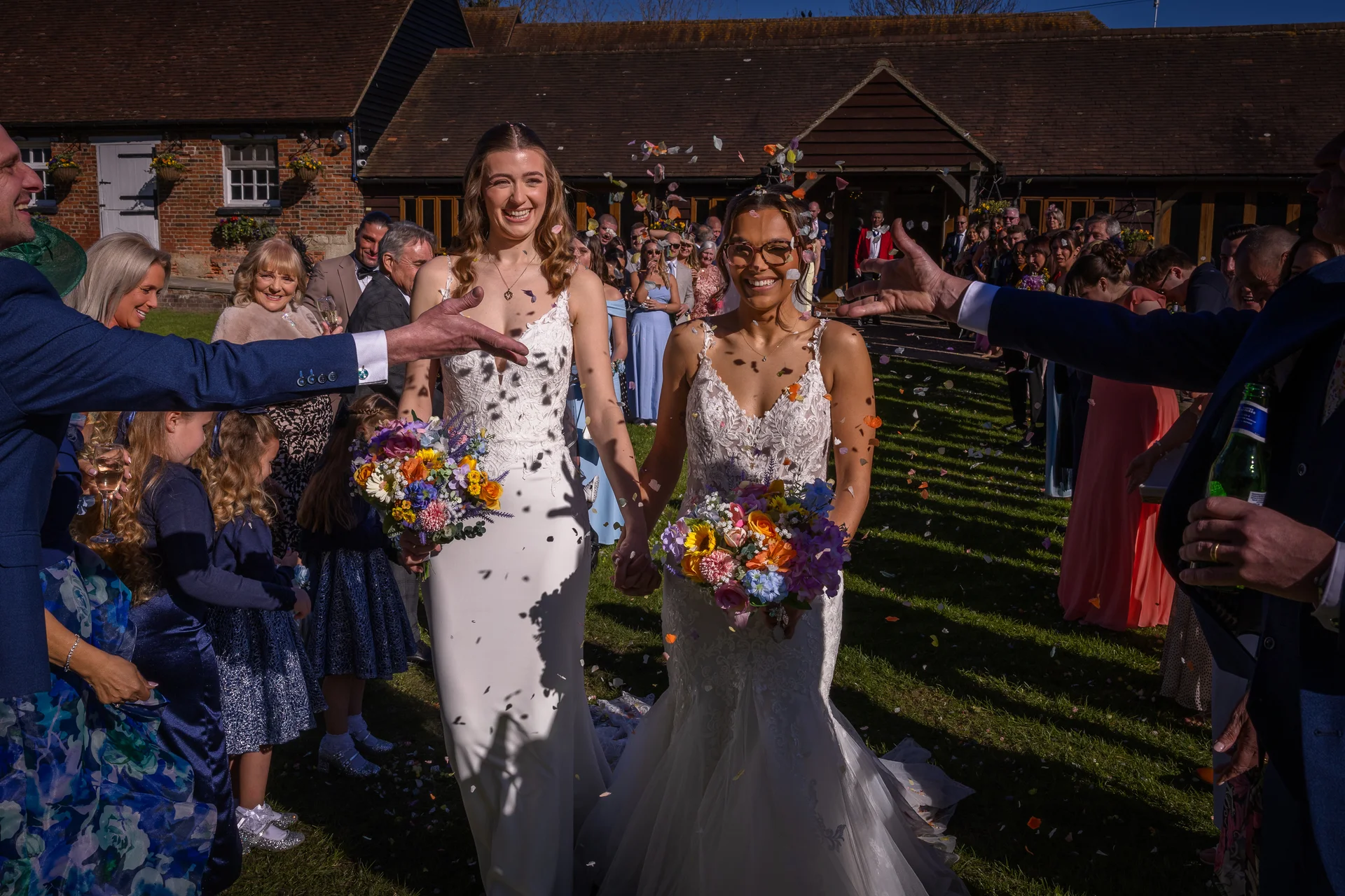 Two smiling brides holding hands and bouquets walk down the aisle as wedding guests shower them with colourful confetti outside a rustic brick barn venue.