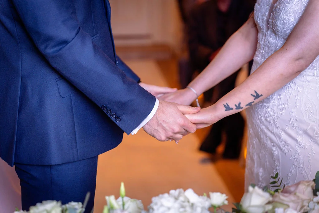 Close-up of a bride and groom holding hands during their ceremony, with the groom wearing a navy suit and the bride in an embellished gown featuring visible arm tattoos.