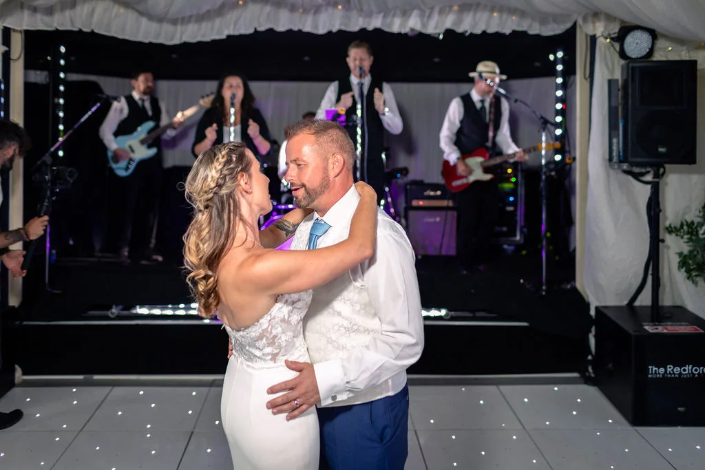 Newlyweds share a romantic first dance on a starlit dance floor in a marquee with a live band performing on stage behind them.