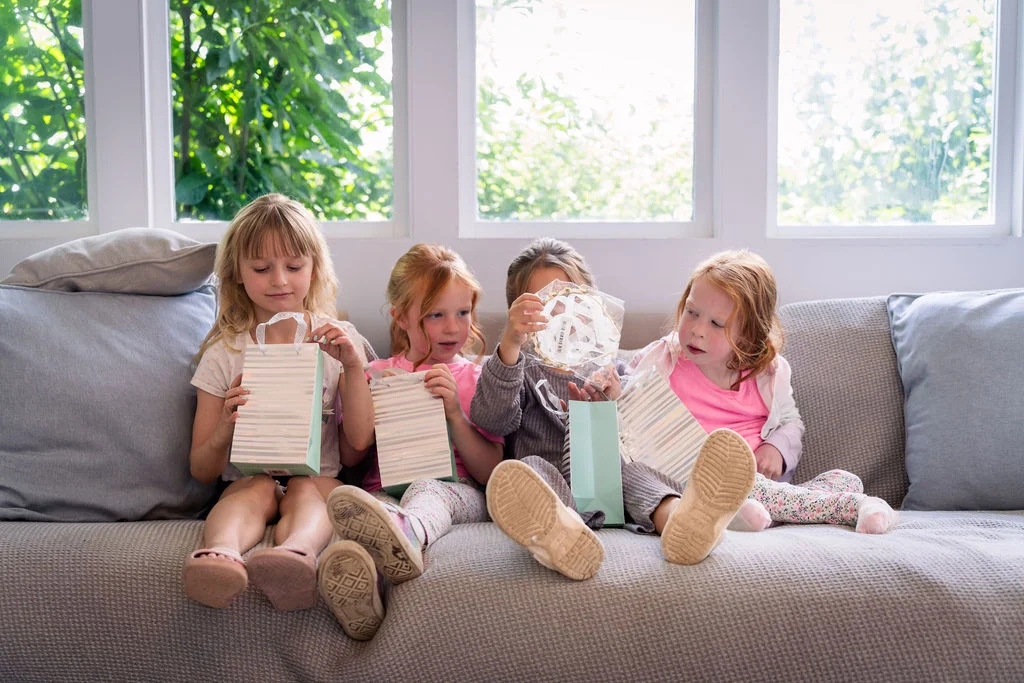 Four young flower girls sit together on a neutral sofa in front of large windows, excitedly opening gift bags during wedding morning preparations.