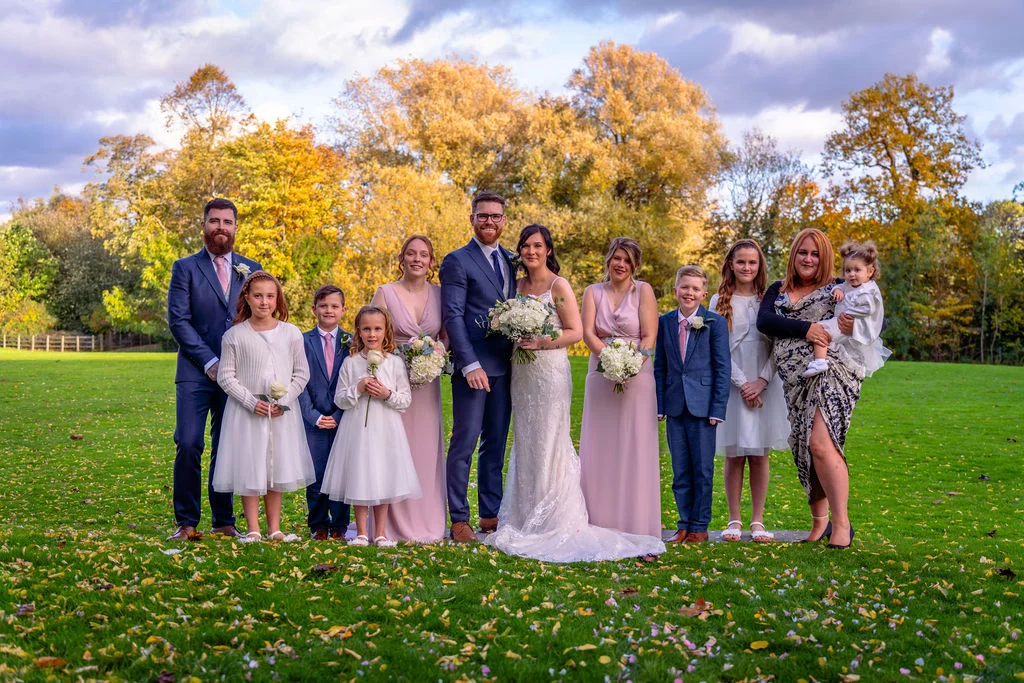 A bridal party and family group pose on a green lawn scattered with fallen autumn leaves against a backdrop of golden trees.
