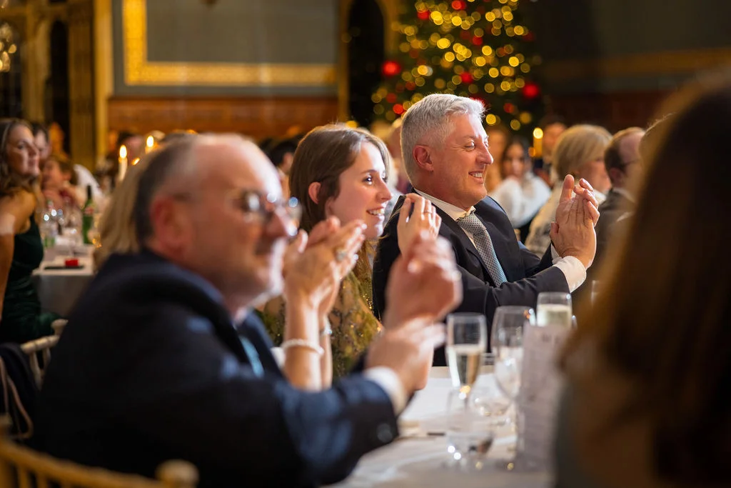 Wedding guests sit at a banquet table indoors during the evening reception, smiling and applauding while a Christmas tree glows in the background.