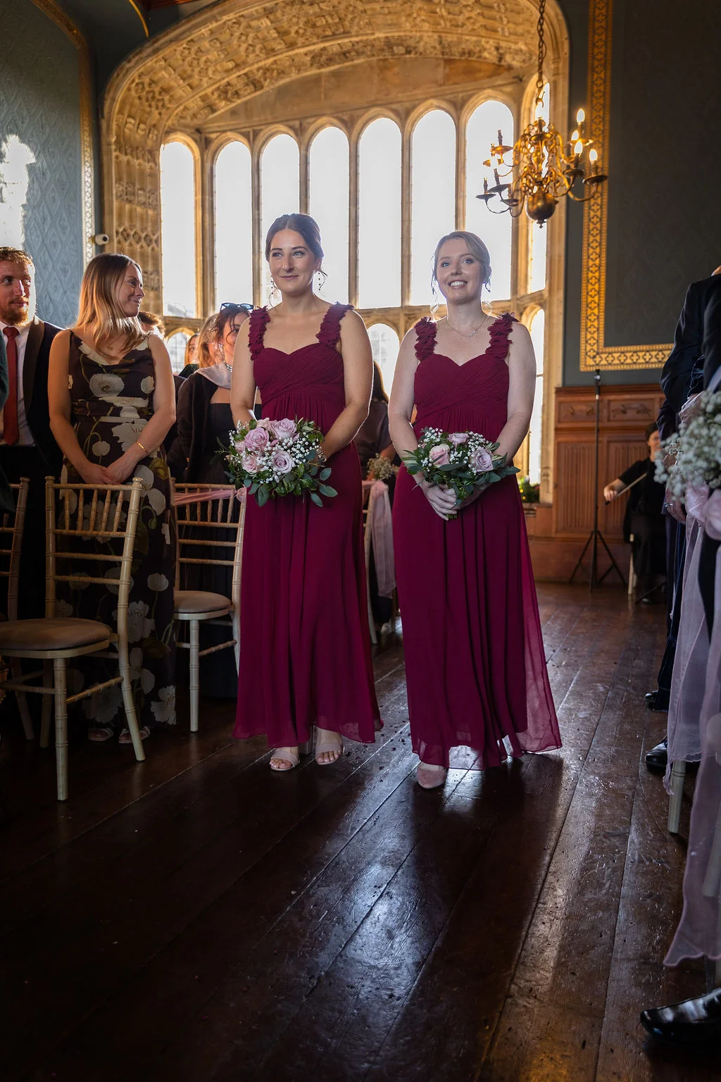 Two smiling bridesmaids in matching burgundy dresses walk down the wooden aisle holding floral bouquets inside a grand hall with arched windows.