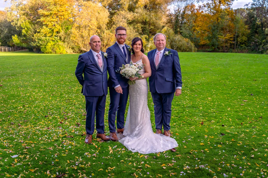 A bride in a lace gown and bouquet stands with the groom and family members on a vibrant green lawn scattered with autumn leaves.