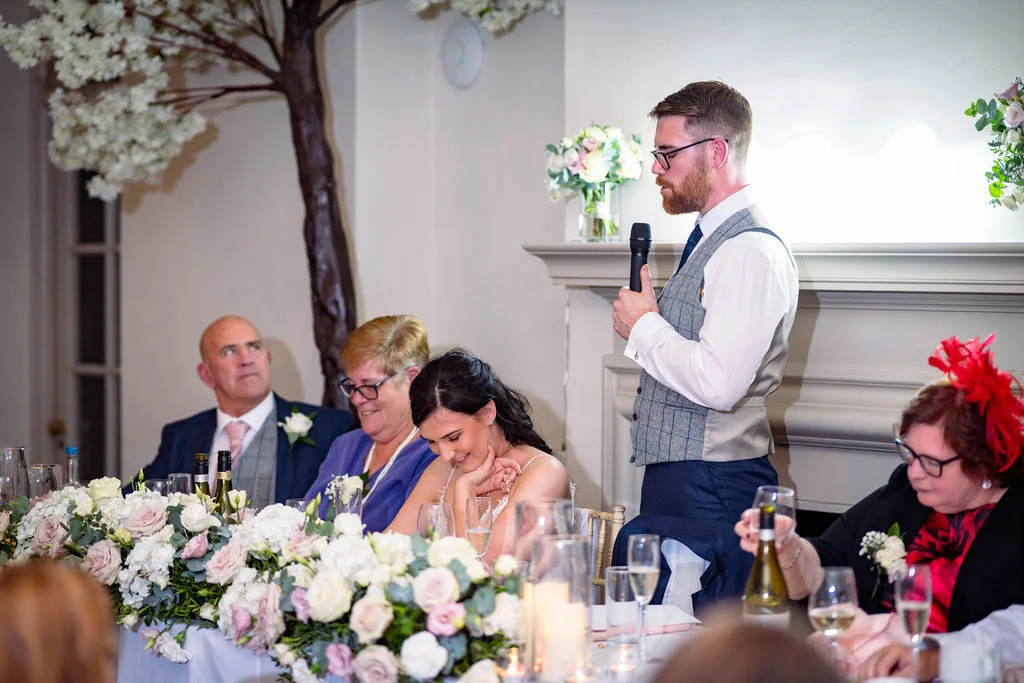 The groom delivers a heartfelt speech holding a microphone while standing near the top table adorned with lush floral centerpieces and candles.