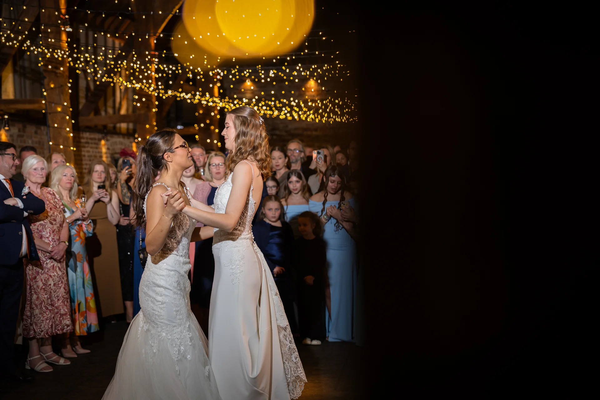 Two brides share their first dance together inside a rustic barn illuminated by a warm fairy light canopy while wedding guests watch.