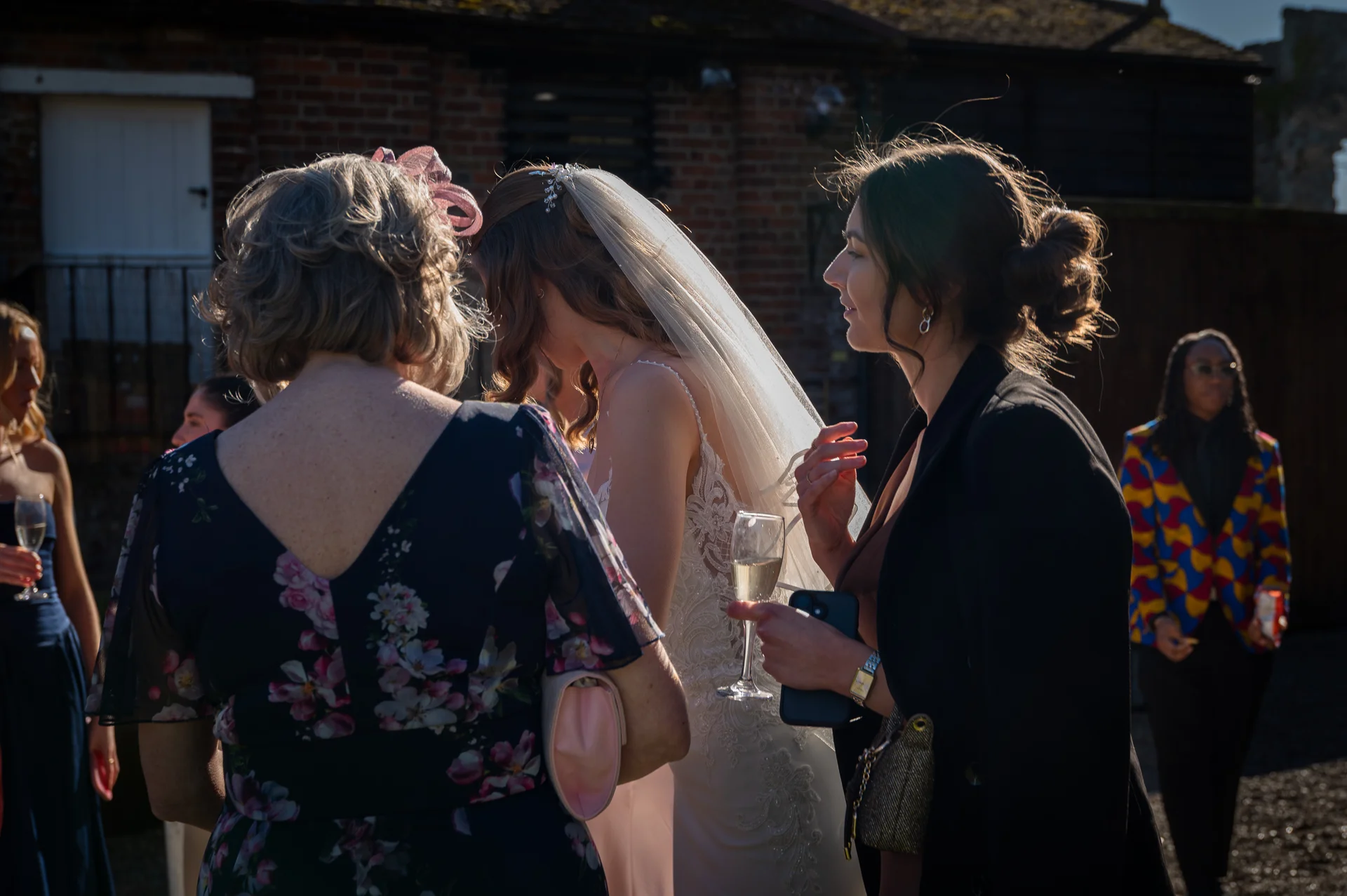 Wedding guests mingle outside in the golden sunlight, holding champagne glasses while chatting near a brick building during the drinks reception.