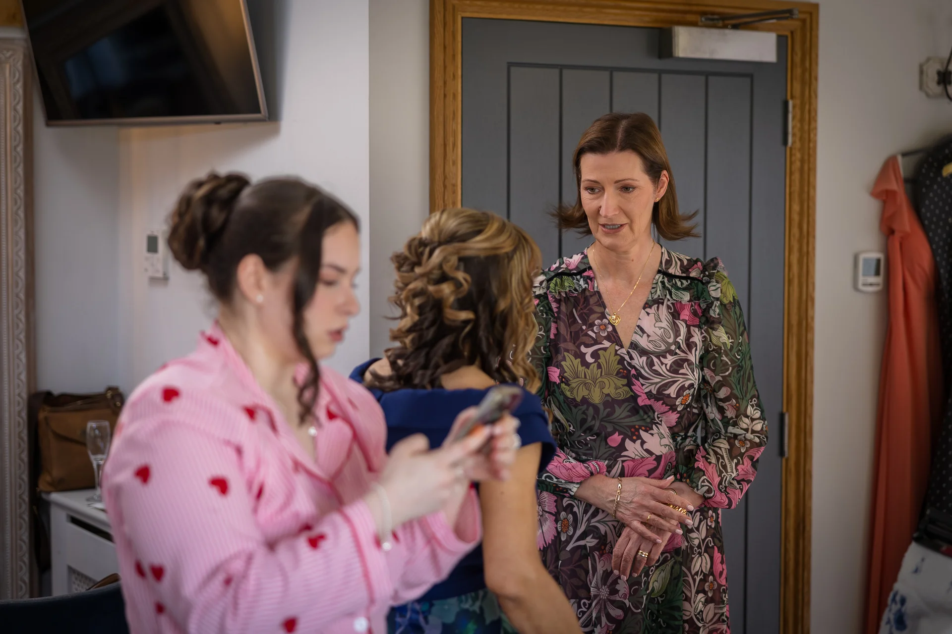 A woman in a floral dress stands talking while a bridesmaid in pink heart-print pyjamas checks her phone during wedding preparations.