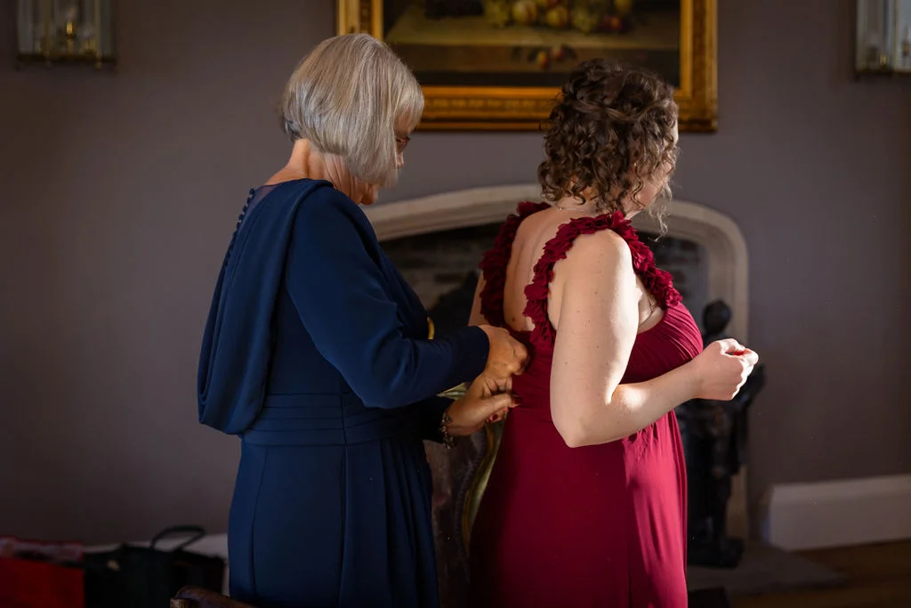 An older woman in a navy dress carefully fastens the back of a red wedding gown for a mature bride with curly hair indoors.