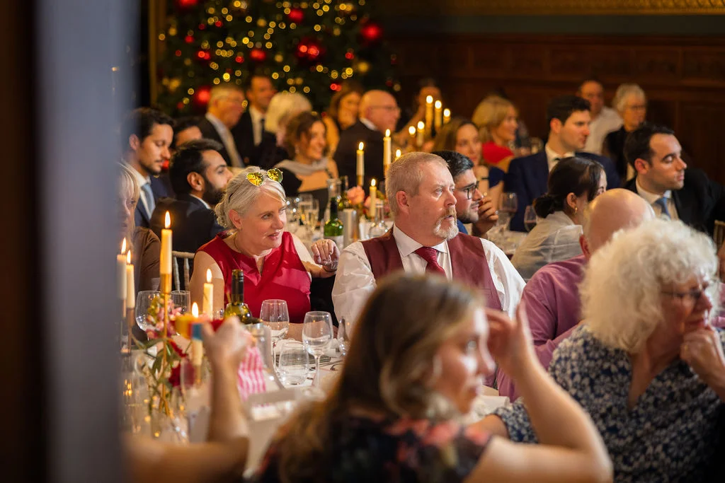 Wedding guests enjoy speeches seated at a long banquet table illuminated by taper candles inside a wood-panelled room with festive decorations.