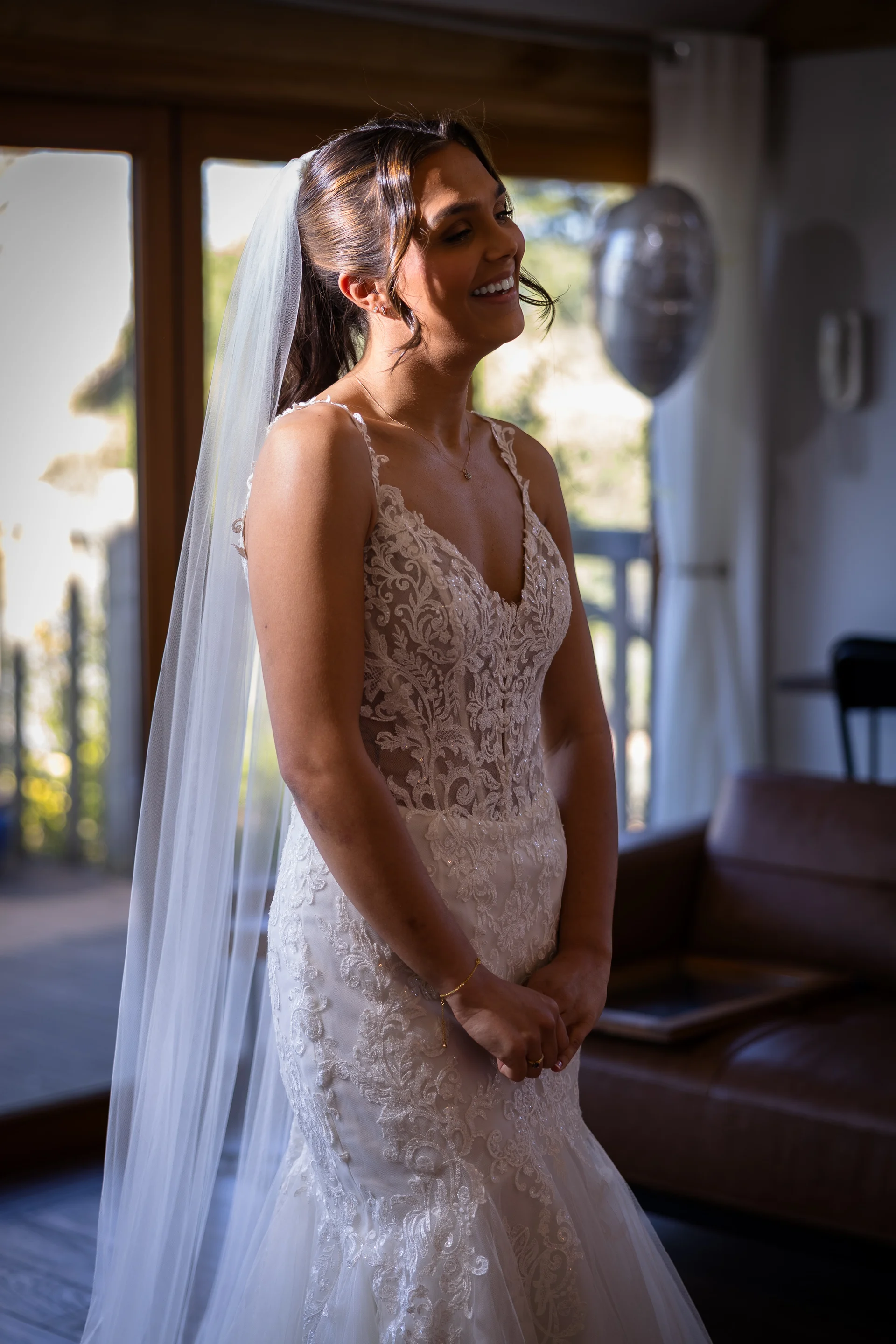 A glowing bride with a high ponytail and lace fit-and-flare gown stands smiling near a window during her preparations.