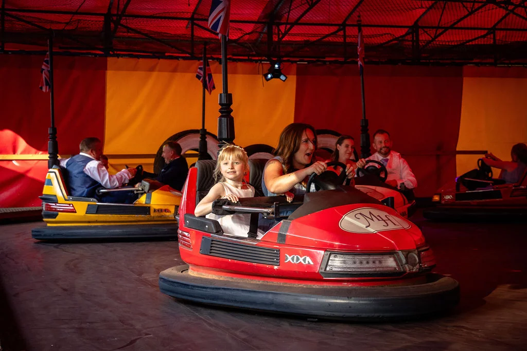 A young flower girl and wedding guests having fun riding bumper cars inside a brightly decorated red and yellow marquee.