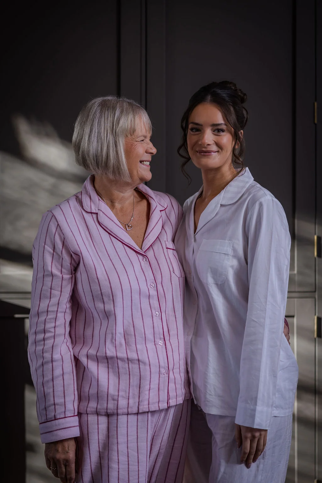 A smiling bride with a sleek low updo wearing white pyjamas poses lovingly next to the mother of the bride in pink striped pyjamas.