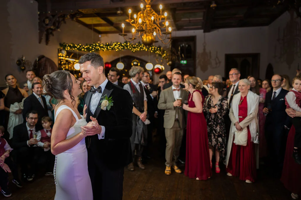 The bride and groom share a joyful first dance in a grand ballroom filled with watching guests under a large golden chandelier and warm fairy lights.