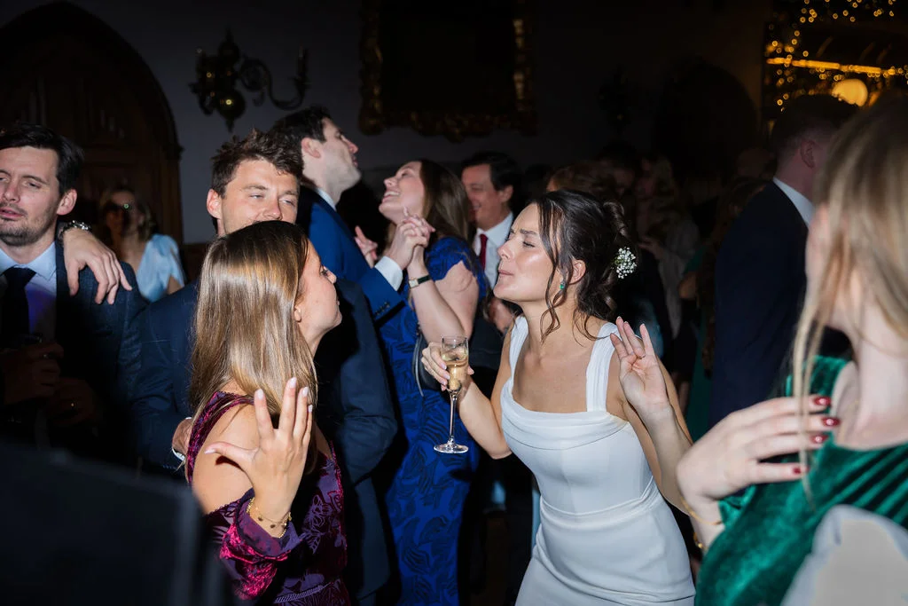 A joyful group of wedding guests dancing and celebrating together during the evening reception, with a bride holding a glass of champagne in the foreground.