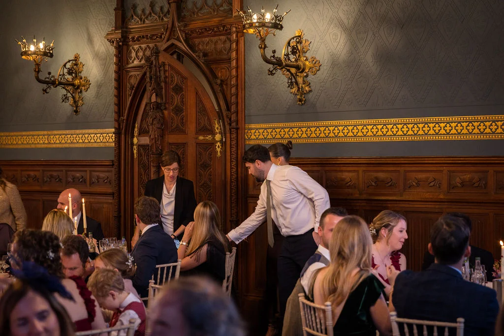 Guests enjoy a wedding breakfast seated at long banquet tables inside an elegant historic hall featuring ornate wooden panelling and decorative wall sconces.