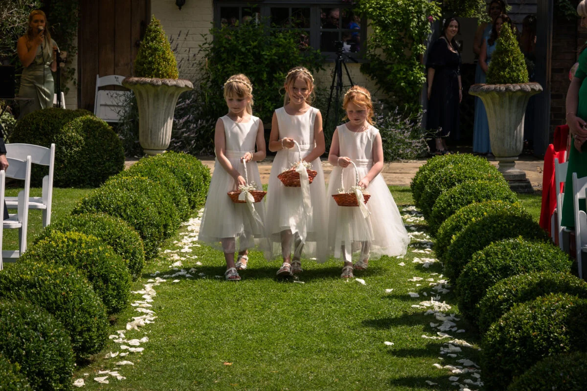 Three young flower girls in white dresses walk down a grass aisle lined with boxwood spheres and white rose petals.