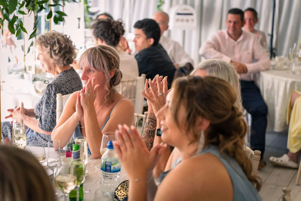 Wedding guests at a reception table laugh and clap joyfully during the speeches, captured in a candid and natural moment.