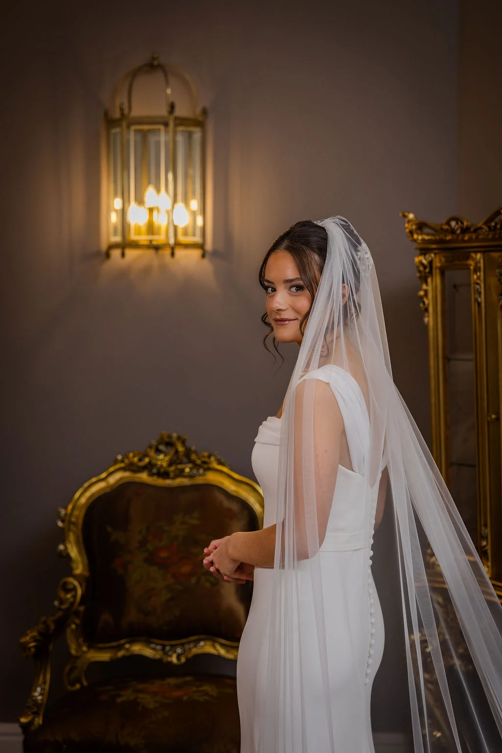 A stunning bride in a sleek satin gown and long chapel veil turns to look back over her shoulder inside an elegant, classically decorated room.