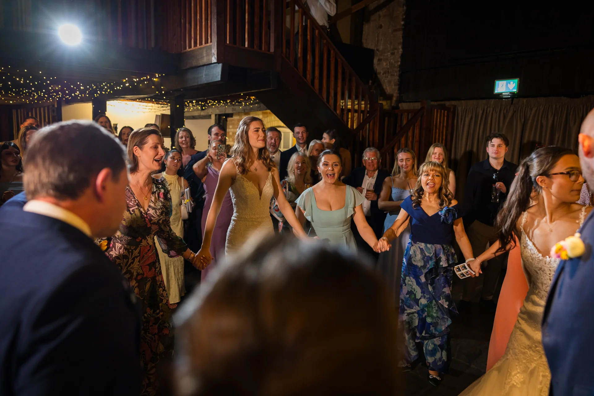 A joyful wedding circle of family and friends hold hands and dance enthusiastically together inside a rustic barn venue illuminated by fairy lights.