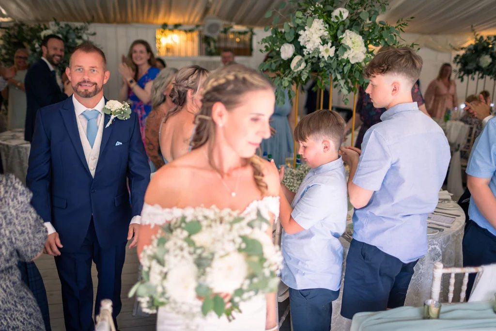 A smiling groom in a navy suit looks on as guests cheer and clap in a marquee wedding reception.