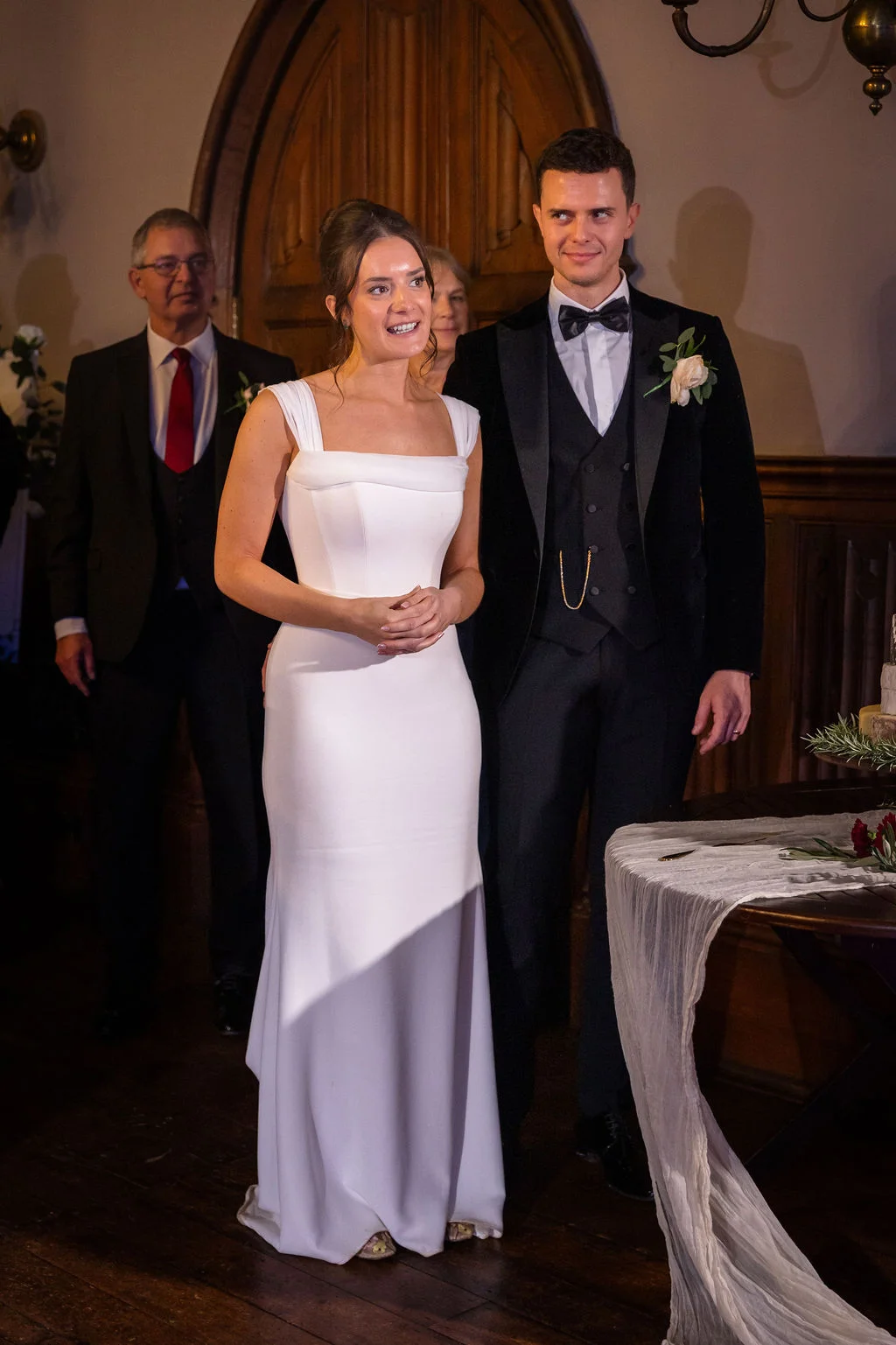 A smiling bride in a modern crepe column gown stands alongside her groom in a velvet tuxedo during an indoor wedding ceremony.